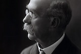 A black and white photo of a bald, moustached man wearing spectacles, suit and tie.