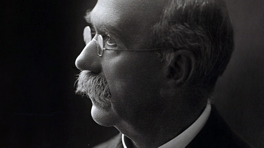 A black and white photo of a bald, moustached man wearing spectacles, suit and tie.