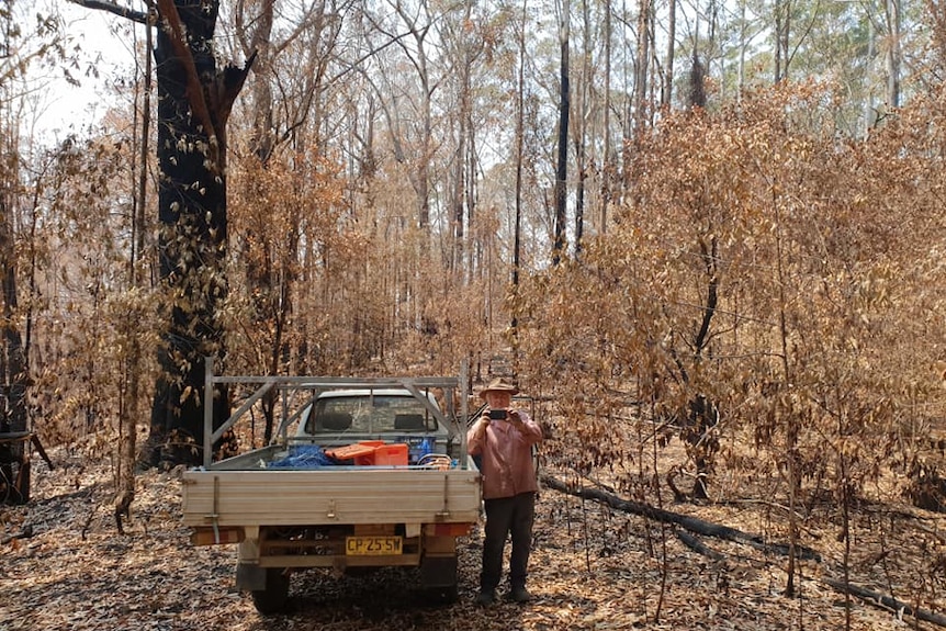 A grey haired man in a hat by his ute standing in scorched yellow bush holding up his phone taking a picture this direction