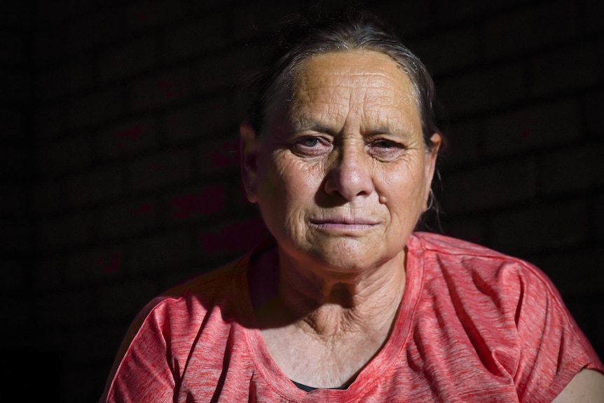 A close up on a woman looking seriously at the camera. She is older, and looks sad. The background is black.