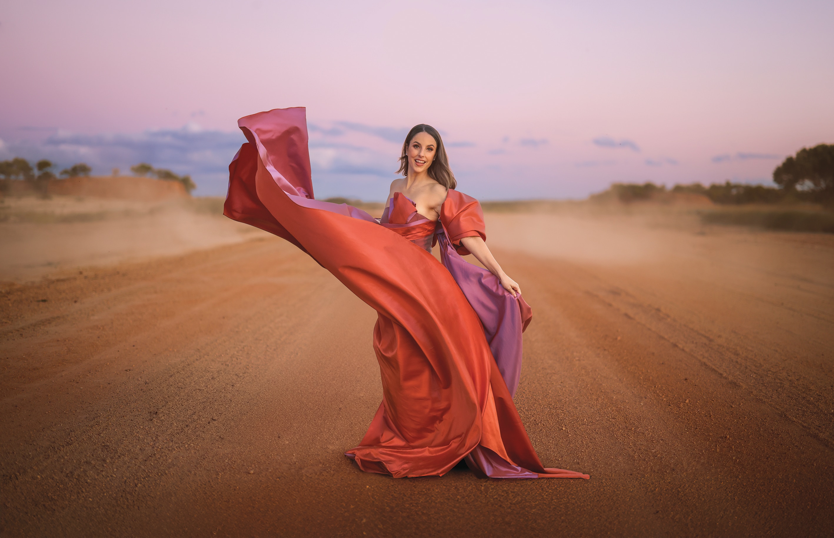 A performer stands on a red dirt outback road at dusk, wearing a flowing red and pink gown that billows in the wind.