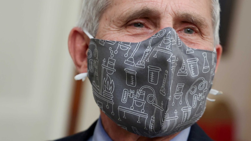 A close up head shot shows an elderly man wearing a grey face mask with cartoon science equipment patterns.