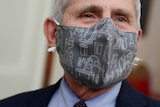 A close up head shot shows an elderly man wearing a grey face mask with cartoon science equipment patterns.