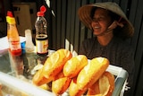 Woman sits behind cabinet in Vietnamese street stall with bred rolls on top of cabinet