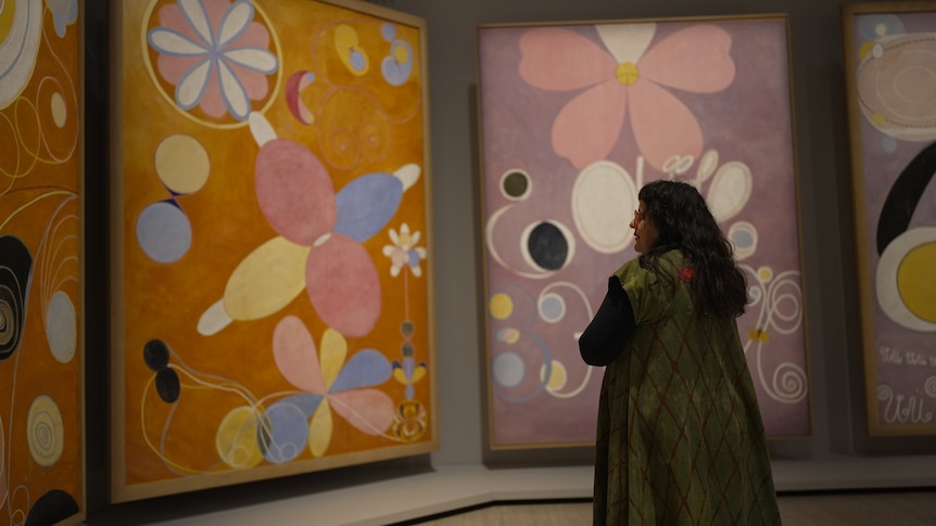 A woman in a green jacket with long dark hair looks at Hilma af Klint’s 3m-high, mauve- and orange-coloured abstract paintings