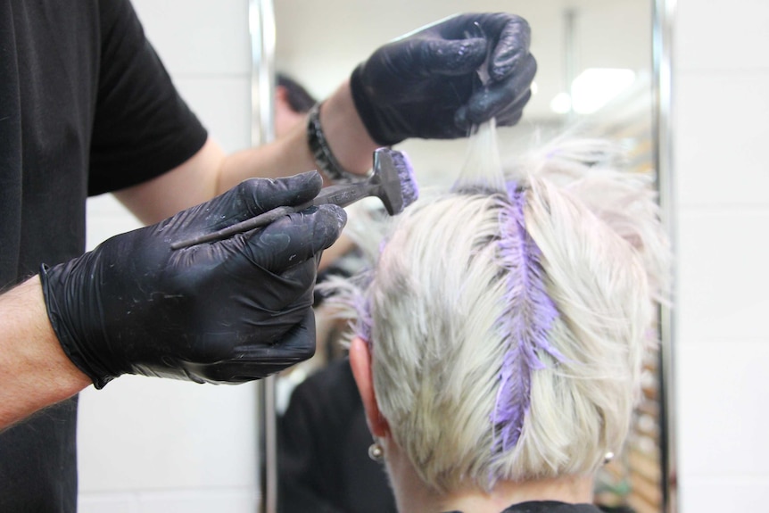 A close up of a hairdresser's hands dying the hair of a woman with blonde hair.