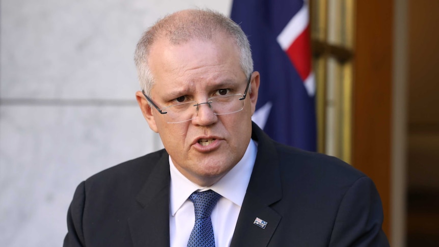 Scott Morrison looks over the top of his glasses as a speaks with an Australian flag behind him