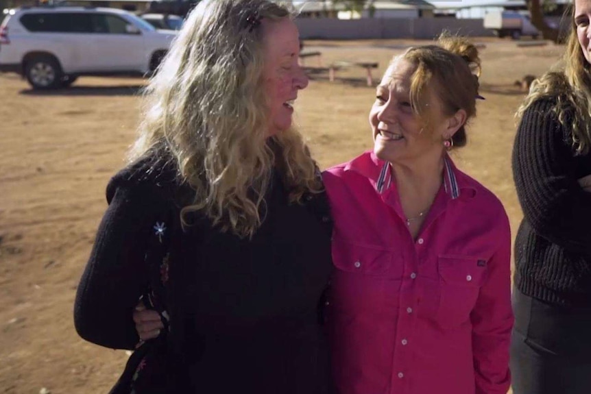 Two women link arms and smile as they talk to one another in the sun.