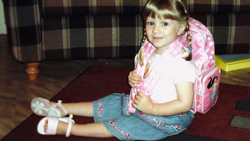 A young smiling schoolgirl with blonde pigtails sits on a red rug. She's wearing a pink tshirt, blue denim shorts and a backpack
