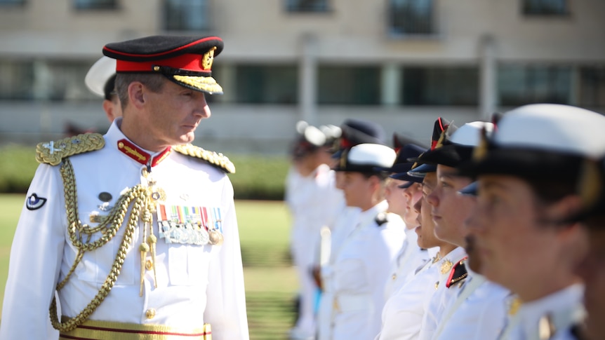 A man in military address walks past younger people in similar dress