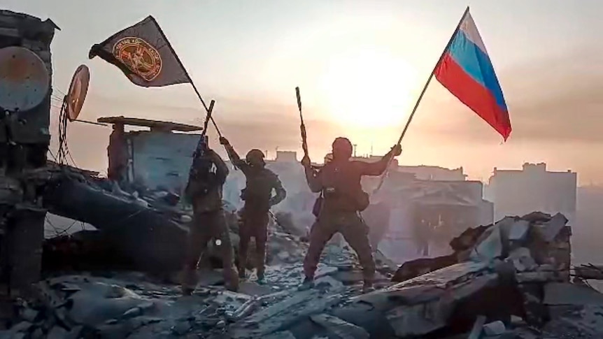 Wagner Group military company members wave a Russian national and Wagner flag atop a damaged building in Bakhmut.