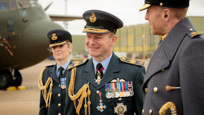 Michael Wigston in full uniform smiles as he looks to the left, alongisde air force colleagues with a military plane in the back