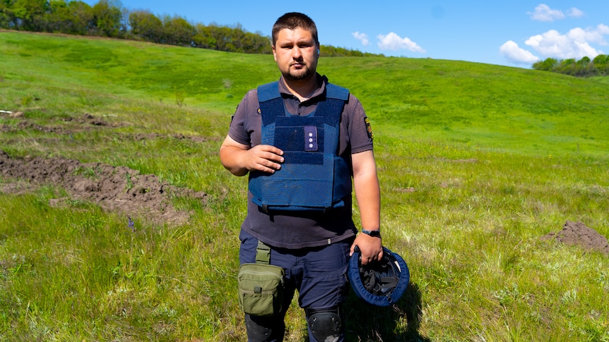 A young bearded man in a bullet proof vest stands in a field holding a helmet