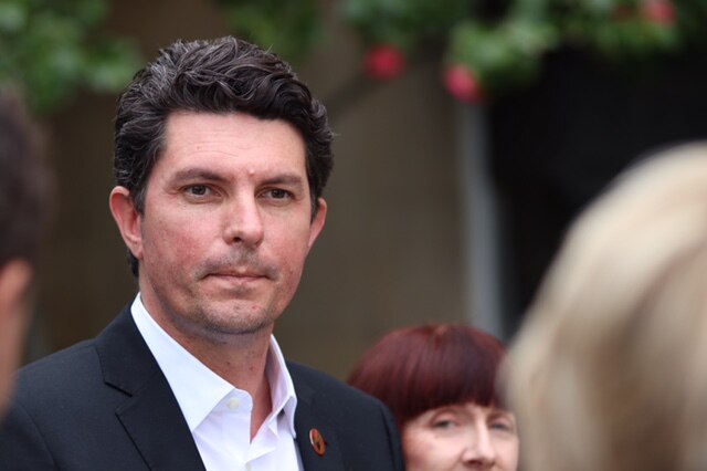 A mid-shot of Scott Ludlam listening to a question from a reporter with part of Rachel Siewert's head behind his left shoulder.