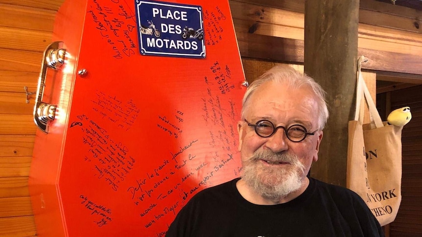 Man with small glasses and moustache stands in front of a red coffin with writing on it