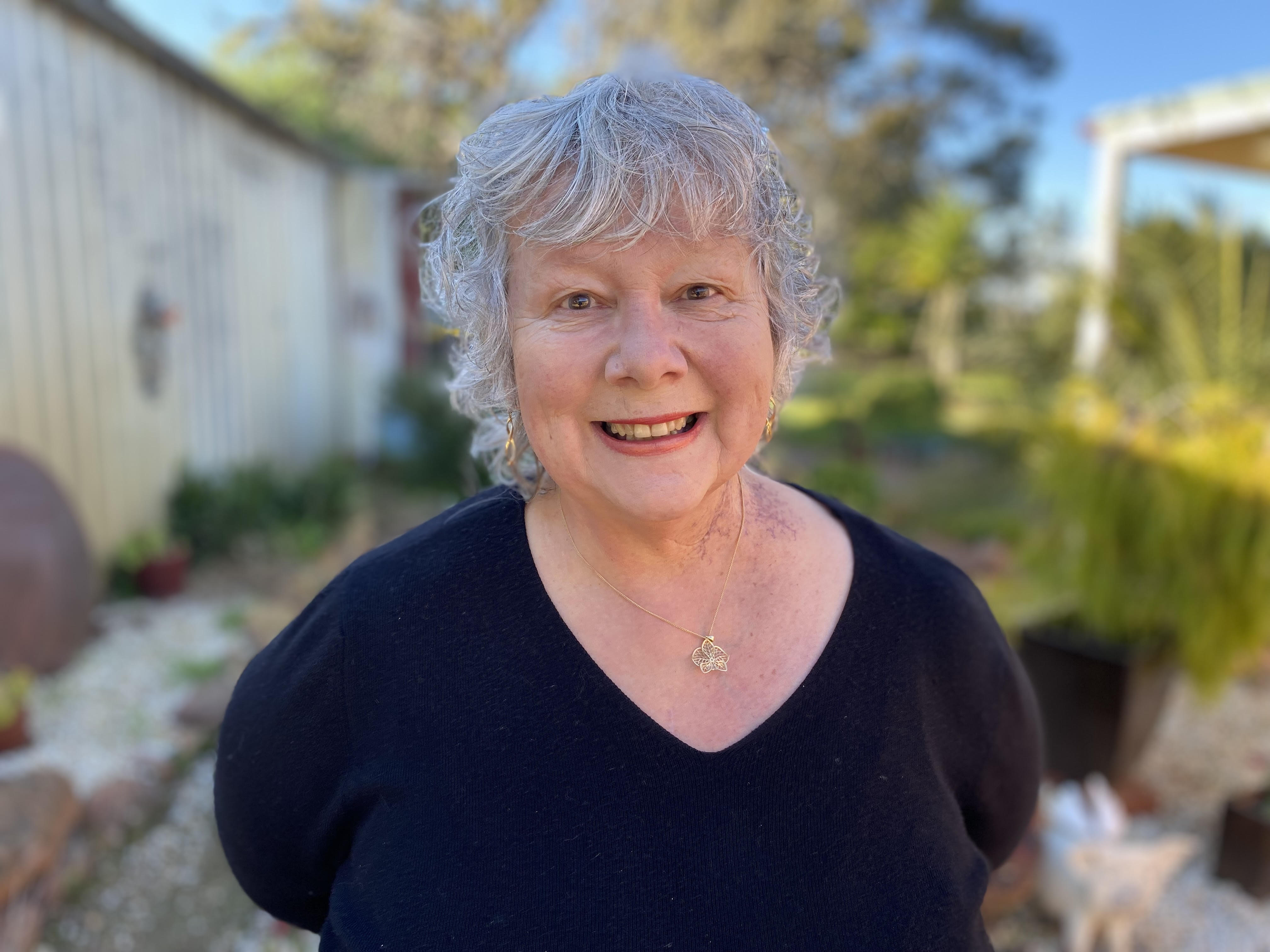 Older, curly grey-haired woman wearing a dark v-necked T-shirt, standing in a back yard