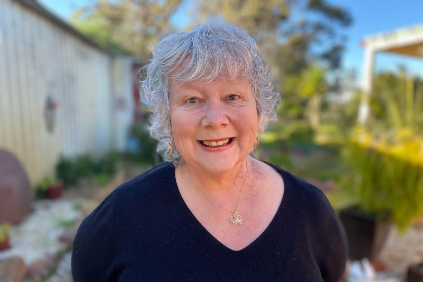 Older, curly grey-haired woman wearing a dark v-necked T-shirt, standing in a back yard