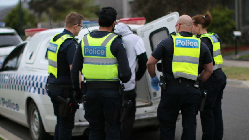 Four police officers watch as an arrested man climbs into the back of a police van.