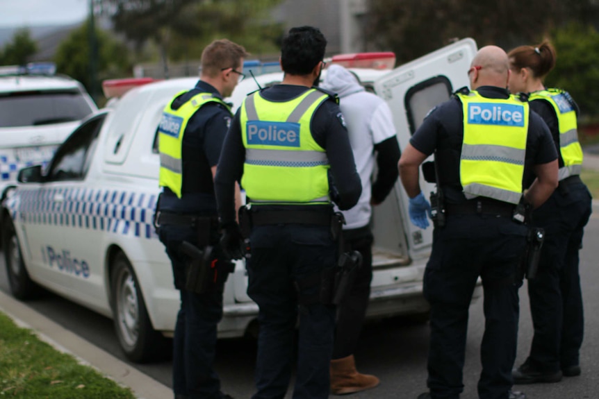 Four police officers watch as an arrested man climbs into the back of a police van.