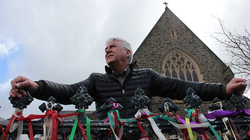 A man stands outside a cathedral in front of a fence covered in coloured ribbons.