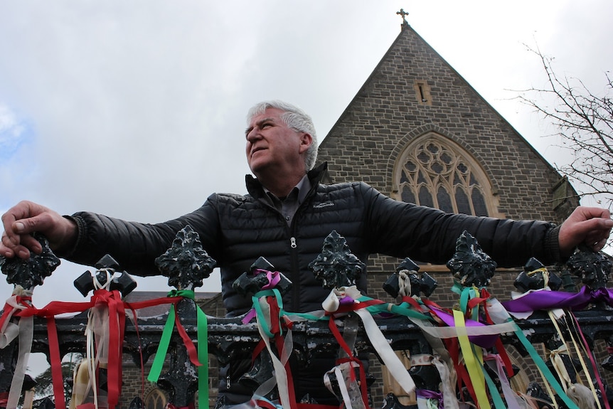 A man stands outside a cathedral in front of a fence covered in coloured ribbons.