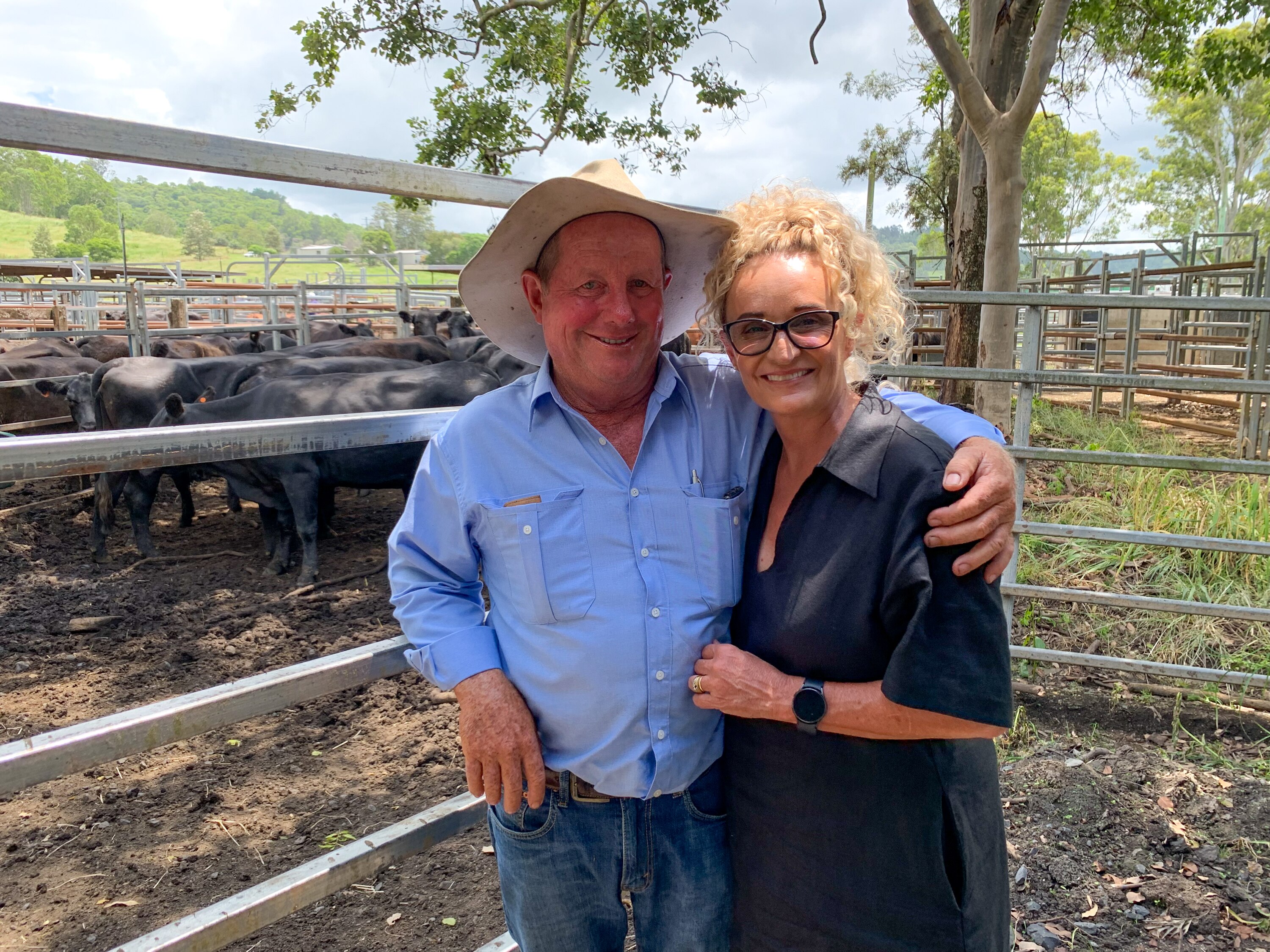A man in a hat and blue shirts stands with his arm around a woman in a black dress in front of black cattle at a saleyard.