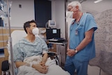 A good-looking man wearing a surgical mask sits up in a hospital bed talking to a white-haired male doctor in blue scrubs.