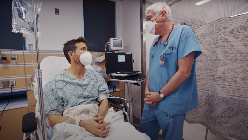 A good-looking man wearing a surgical mask sits up in a hospital bed talking to a white-haired male doctor in blue scrubs.