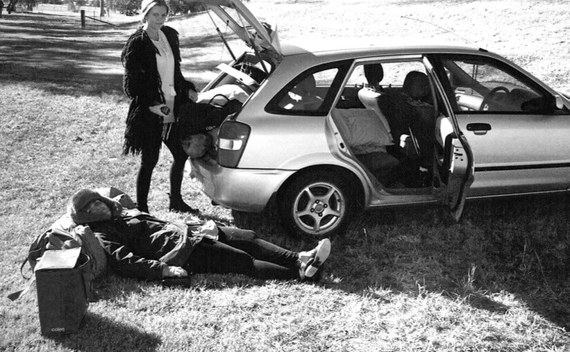 A black and white photo of two friends packing a hatchback car. One likes on bags on teh ground, the other stands by the boot