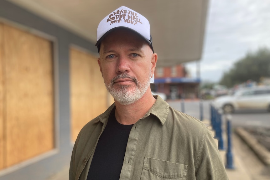 A man wearing a cap that reads 'Where the muddy hell are you?' Boarded up windows are in the background.