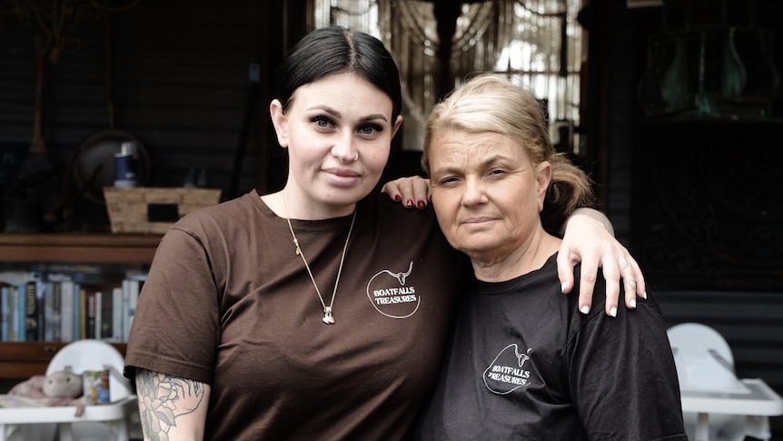 A serious older woman, slightly smiling younger woman, tattoos on arm, stand arm in arm, both wear brown tee with logo. 