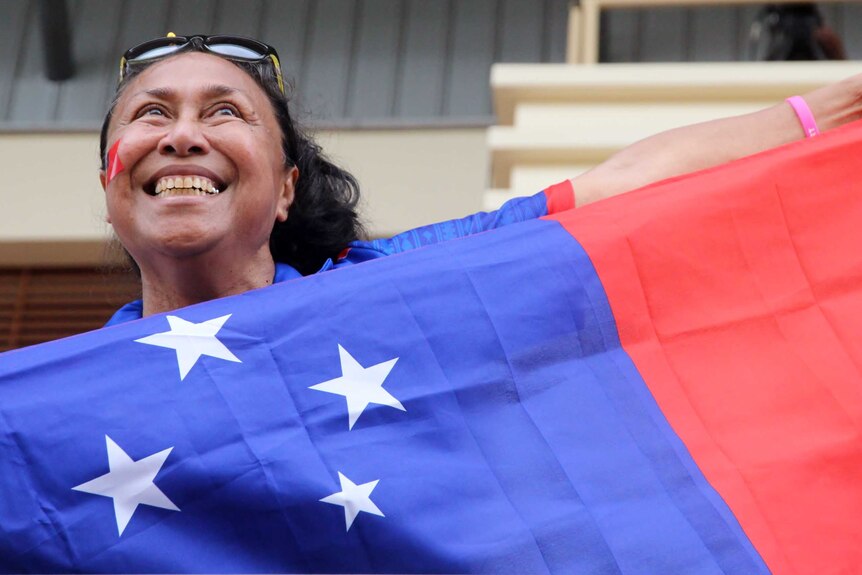 A dark-haired, smiling Samoan woman holds the red, white and blue flag of Samoan under her chin while cheering in a Grandstand.