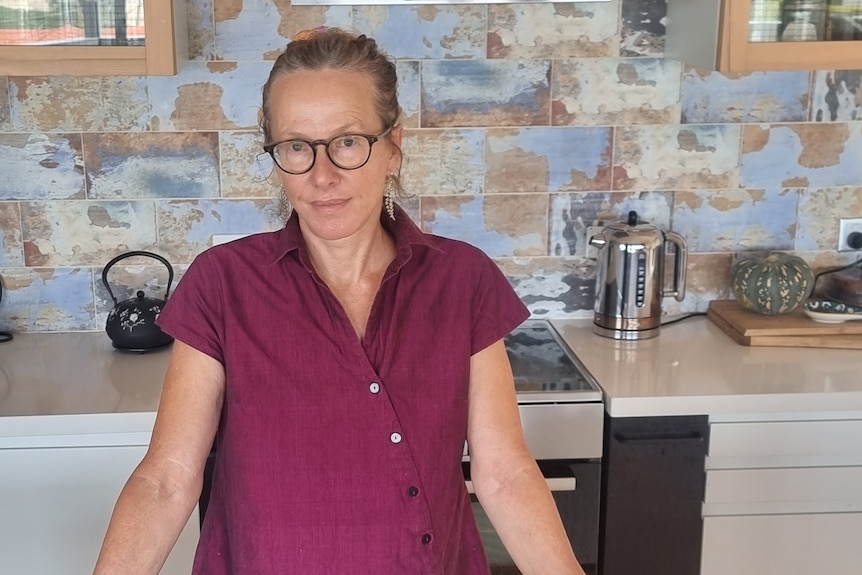 A woman wearing glasses and a red blouse stands in her kitchen.