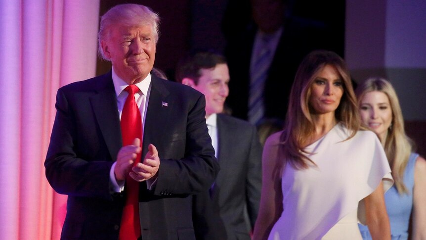 Donald Trump smiles and claps as Melania stands behind him looking concerned.