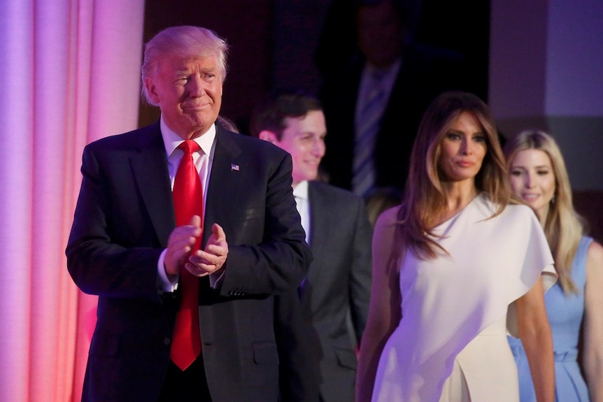 Donald Trump smiles and claps as Melania stands behind him looking concerned.