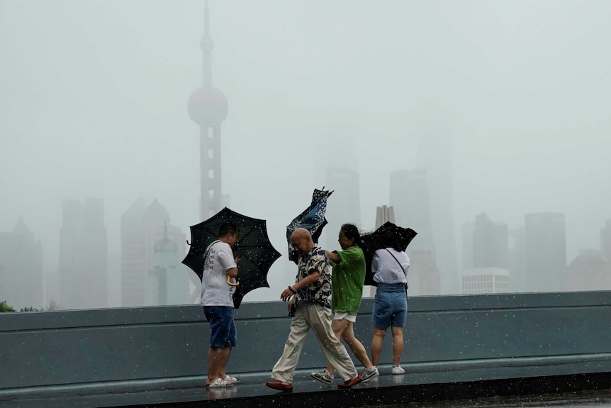 Looking across a road, you view four people in front of a silhouette of Shanghai's skyline as they struggle to open umbrellas.