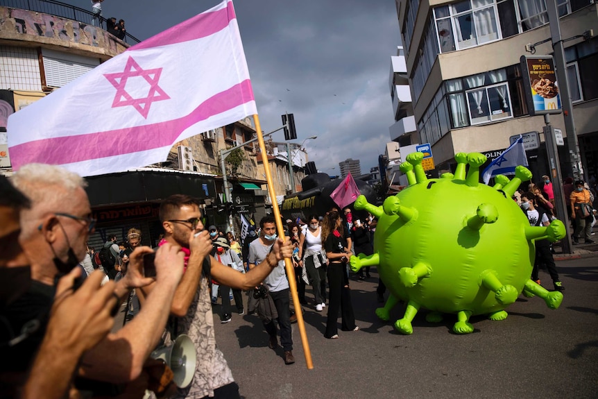 A crowd carries a pick Israeli flag and an inflatable model of a coronavirus.
