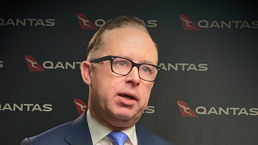 Alan Joyce seated, gesturing his hands, with Qantas logos behind him.