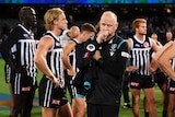 An AFL coach covers his mouth as his dejected players stand in the background.
