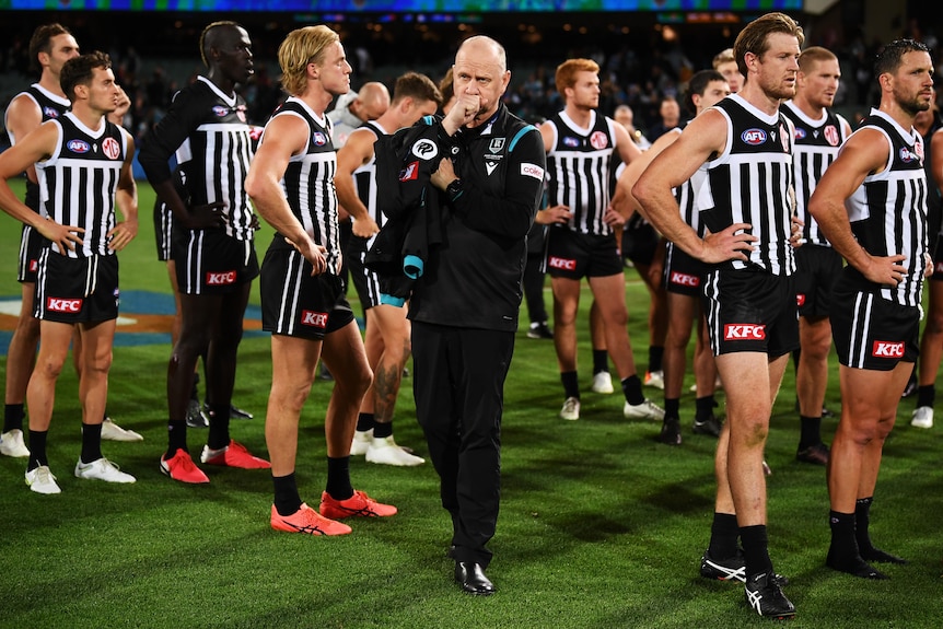 An AFL coach covers his mouth as his dejected players stand in the background.