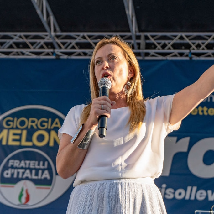 A blonde woman in white speaks into a microphone with her left hand raised