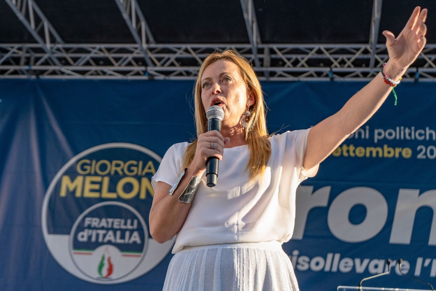 A blonde woman in white speaks into a microphone with her left hand raised