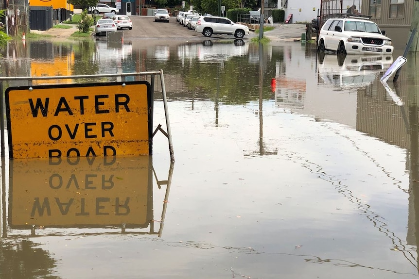 A sign that says water over road is half covered by water and a car is also partly underwater