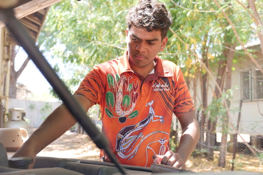 A young Indigenous man wears a bright orange T-shirt while fixing a car