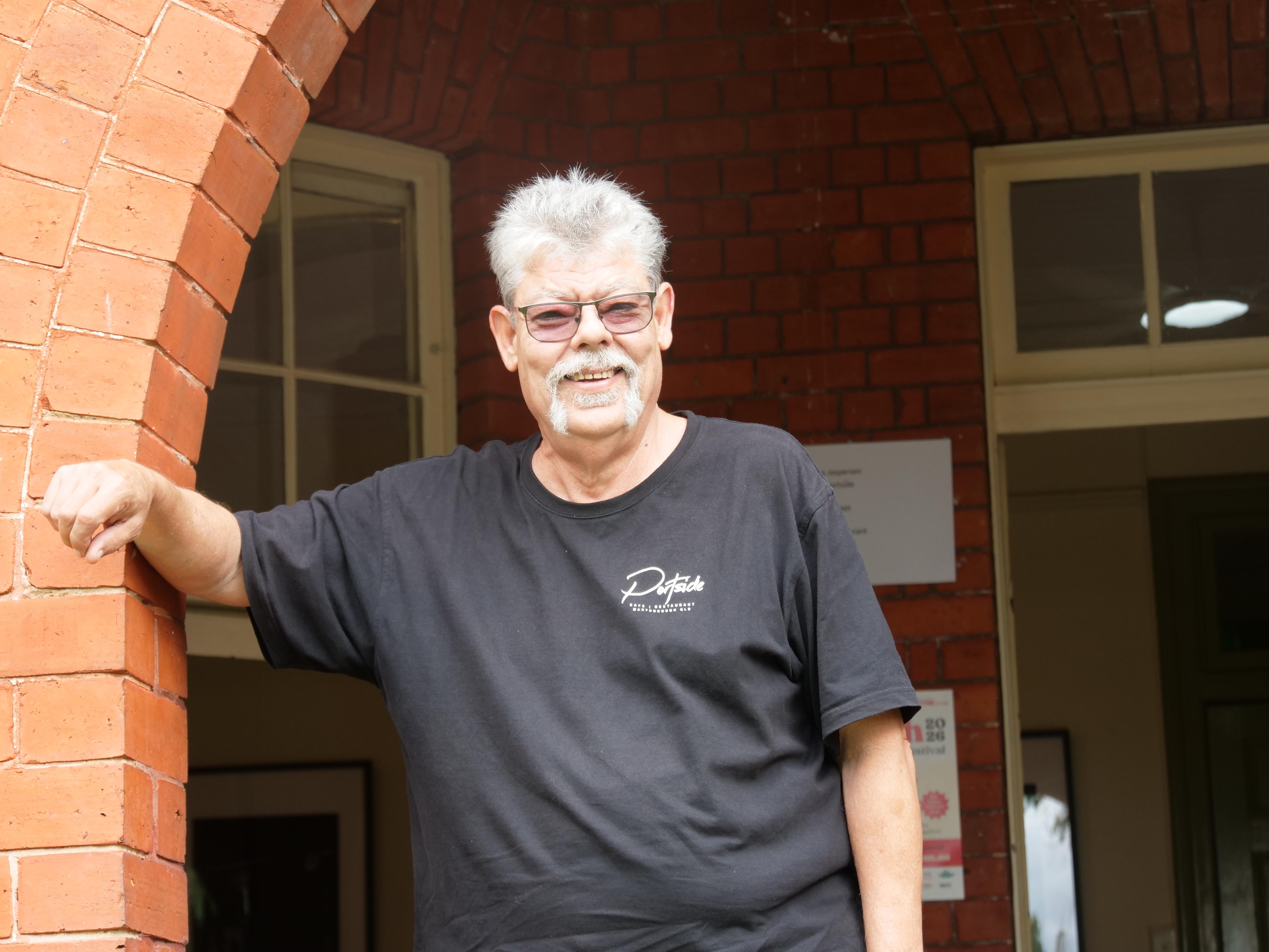 A man with dark framed glasses smiles while leaning against a brick alcove.