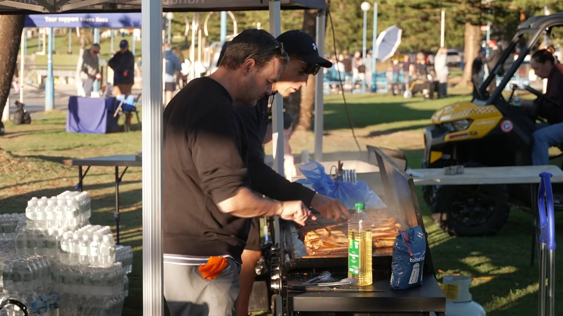 Two men use tongs to turn sausages on a bbq in a park.