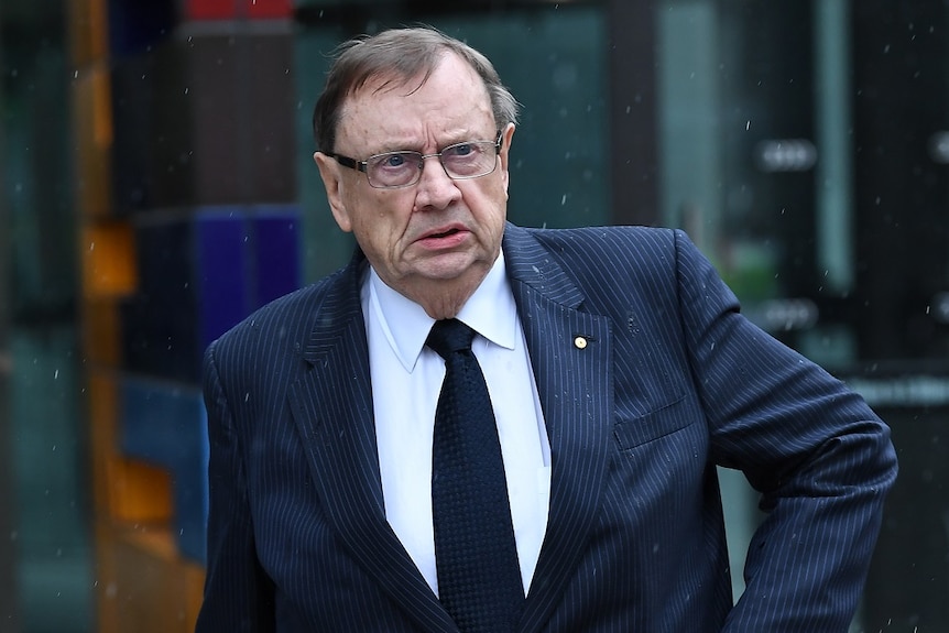 Harold Mitchell walks out of glass doors of the court building into light rain while wearing a suit.