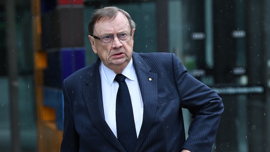 Harold Mitchell walks out of glass doors of the court building into light rain while wearing a suit.