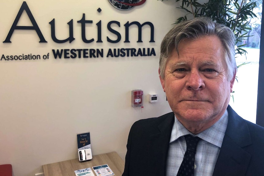 A head and shoulders shot of a smiling Russell Thomas standing in an office with Autism Association of WA written on the wall.