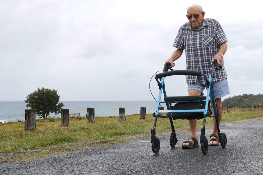 A man walks along the road by the seaside using a walker on wheels for support.